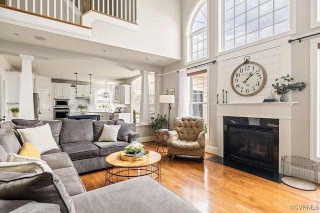 living room featuring ornate columns, a towering ceiling, light wood-style flooring, a fireplace with flush hearth, and baseboards