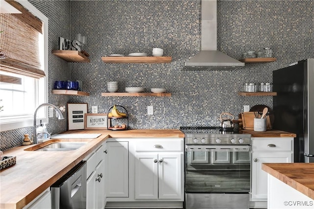 kitchen with butcher block counters, a sink, wall chimney range hood, appliances with stainless steel finishes, and open shelves