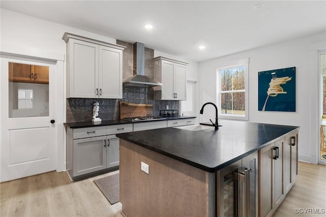 kitchen with stainless steel gas cooktop, dark countertops, a sink, beverage cooler, and wall chimney exhaust hood