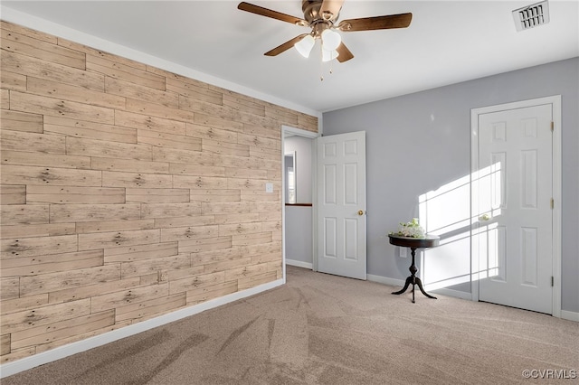 carpeted empty room with visible vents, wood walls, baseboards, and ceiling fan