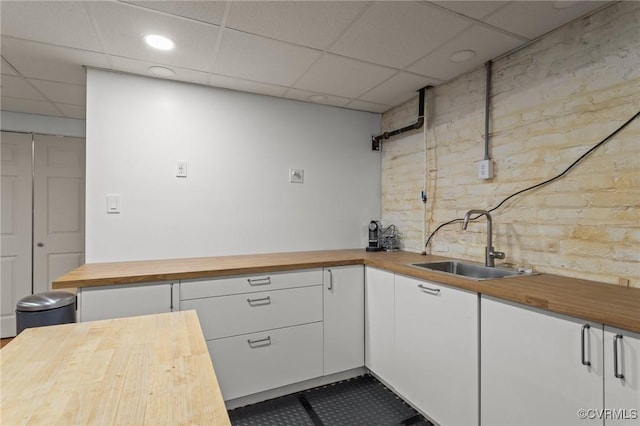 kitchen with butcher block countertops, a sink, white cabinetry, brick wall, and a paneled ceiling