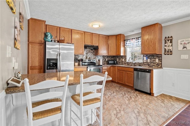 kitchen with under cabinet range hood, tasteful backsplash, stainless steel appliances, a peninsula, and brown cabinetry