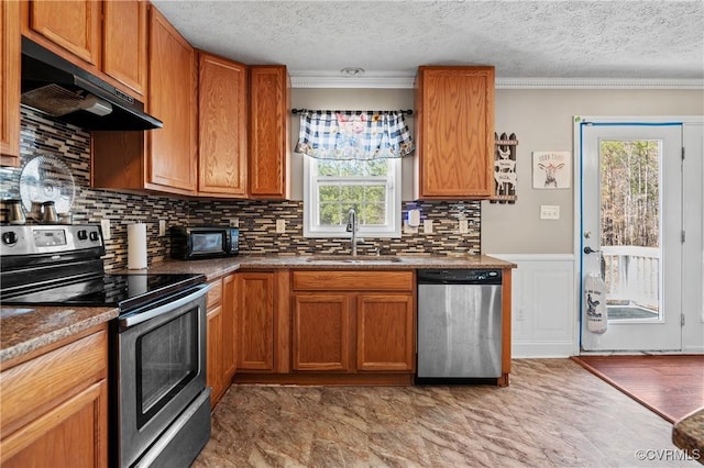 kitchen featuring brown cabinets, under cabinet range hood, a sink, appliances with stainless steel finishes, and crown molding