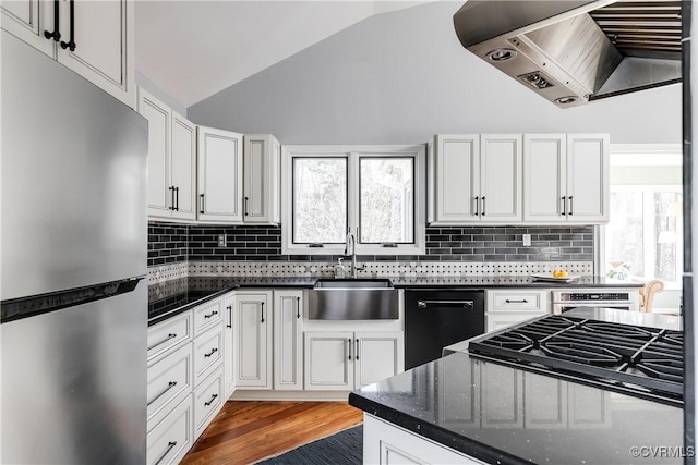 kitchen featuring lofted ceiling, appliances with stainless steel finishes, white cabinetry, a sink, and wall chimney range hood