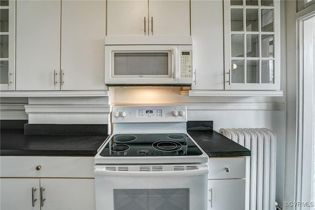 kitchen with white appliances, radiator heating unit, white cabinets, glass insert cabinets, and dark countertops