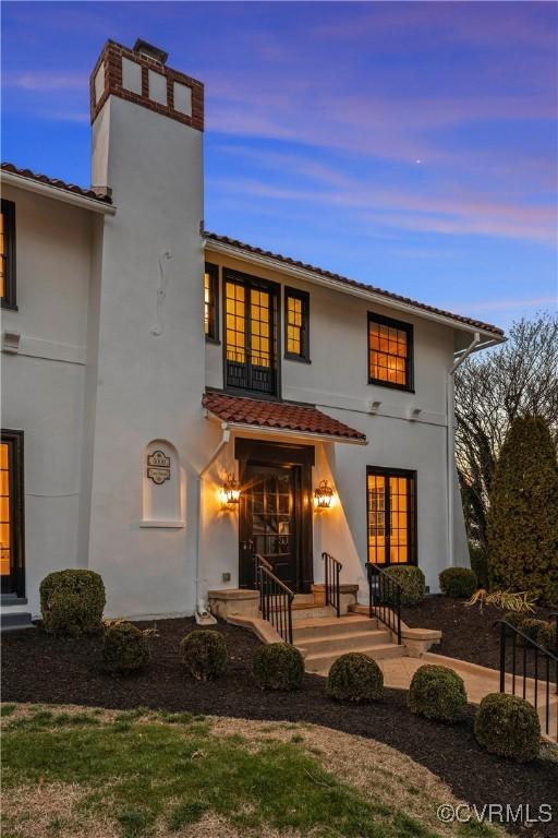 back of house at dusk featuring stucco siding, french doors, and a chimney