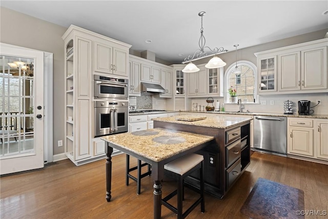 kitchen with under cabinet range hood, stainless steel appliances, dark wood-style flooring, a center island, and decorative backsplash