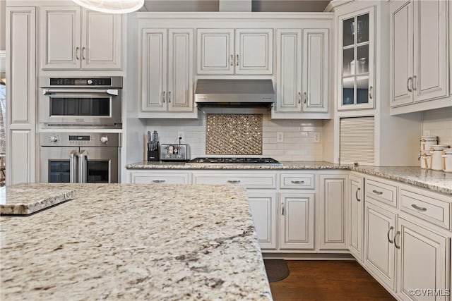 kitchen with dark wood finished floors, decorative backsplash, glass insert cabinets, stainless steel double oven, and under cabinet range hood