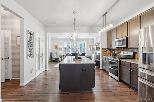 kitchen with light stone counters, dark wood-style flooring, a kitchen island with sink, decorative backsplash, and appliances with stainless steel finishes