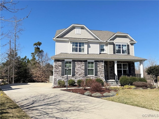 view of front of property with roof with shingles, covered porch, concrete driveway, a front yard, and stone siding