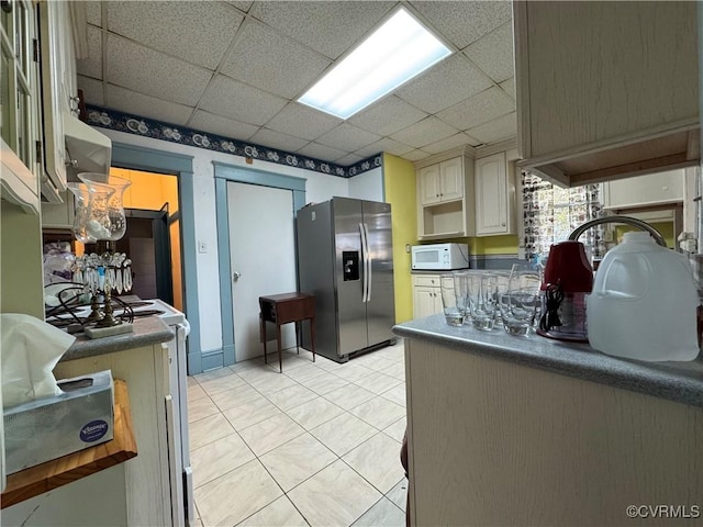 kitchen with light tile patterned floors, a drop ceiling, dark countertops, white microwave, and stainless steel refrigerator with ice dispenser