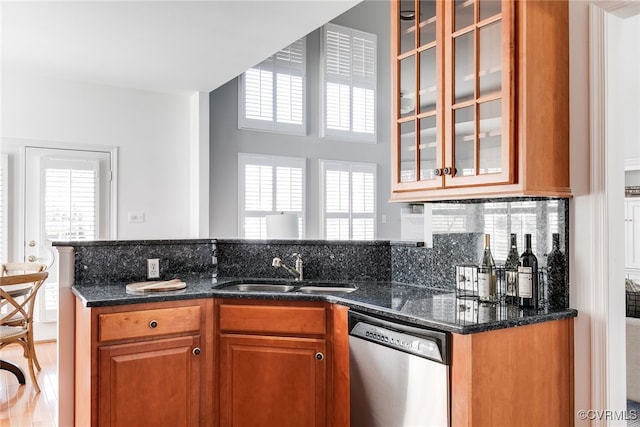 kitchen featuring a sink, stainless steel dishwasher, backsplash, brown cabinetry, and glass insert cabinets