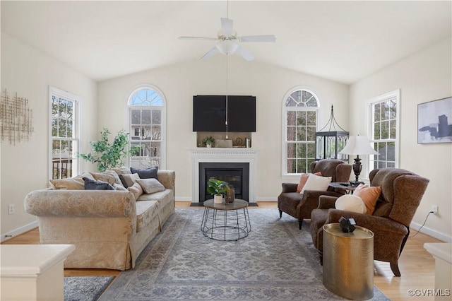 living area with a ceiling fan, a glass covered fireplace, vaulted ceiling, light wood-type flooring, and baseboards