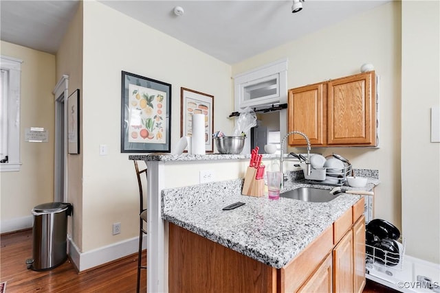 kitchen featuring baseboards, brown cabinetry, light stone counters, dark wood-type flooring, and a sink