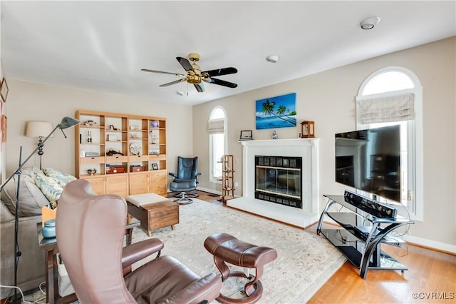 living room with a glass covered fireplace, wood finished floors, baseboards, and a ceiling fan