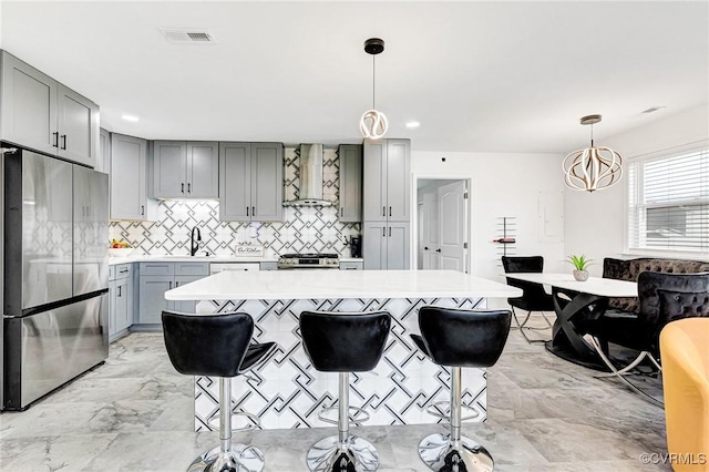 kitchen with marble finish floor, gray cabinets, stainless steel refrigerator, wall chimney exhaust hood, and light countertops