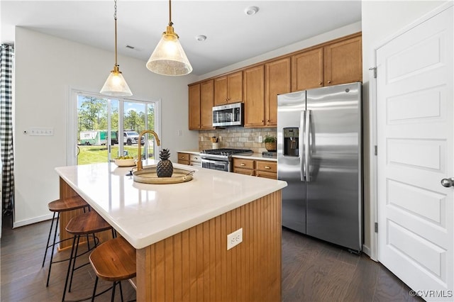 kitchen with a center island with sink, decorative backsplash, appliances with stainless steel finishes, brown cabinets, and dark wood-style flooring