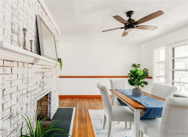 dining space with baseboards, a ceiling fan, wood finished floors, a textured ceiling, and a brick fireplace