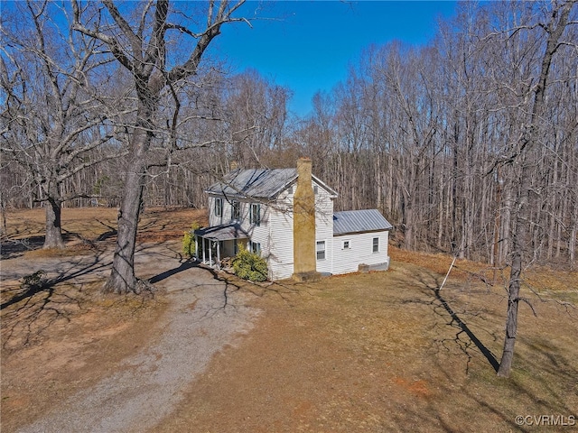 view of side of property with dirt driveway, metal roof, a chimney, and a view of trees