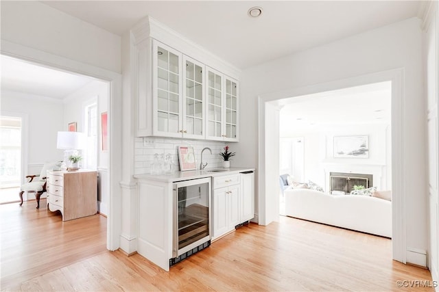 bar featuring light wood-type flooring, beverage cooler, a sink, a glass covered fireplace, and decorative backsplash