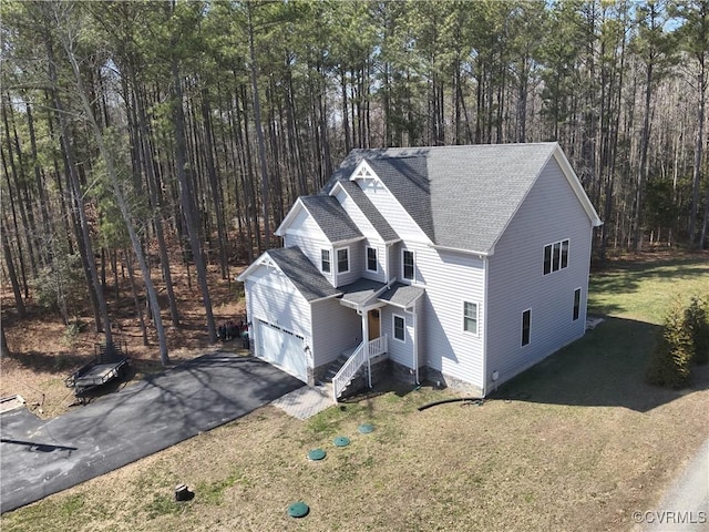 view of front facade featuring roof with shingles, an attached garage, a wooded view, driveway, and a front lawn