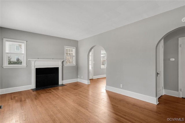 unfurnished living room with a fireplace with flush hearth, arched walkways, light wood-type flooring, and baseboards