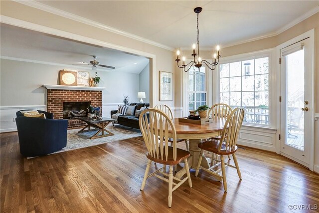 dining space featuring a wainscoted wall, ceiling fan with notable chandelier, ornamental molding, a brick fireplace, and dark wood finished floors