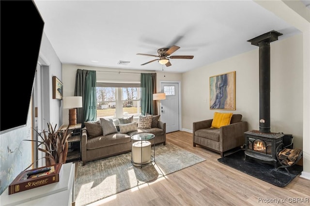 living room with ceiling fan, wood finished floors, visible vents, baseboards, and a wood stove