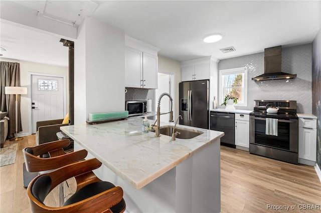 kitchen with light wood finished floors, stainless steel appliances, wall chimney range hood, white cabinetry, and a sink