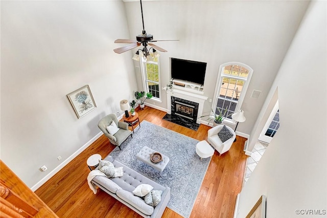 living room with ceiling fan, wood finished floors, a high end fireplace, baseboards, and a towering ceiling