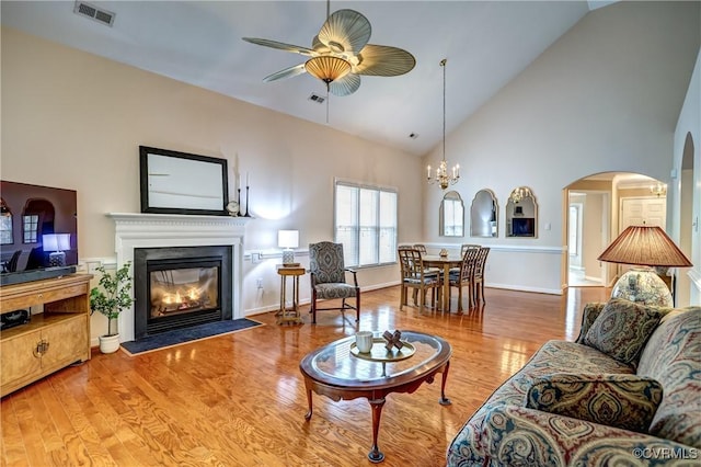 living room with visible vents, a glass covered fireplace, wood finished floors, high vaulted ceiling, and ceiling fan with notable chandelier