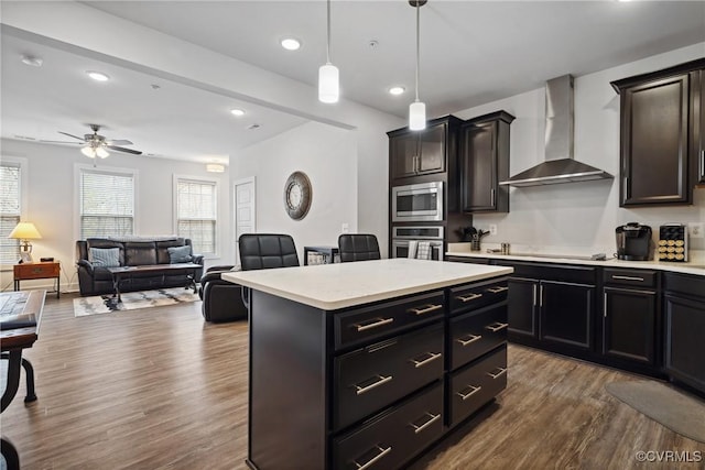 kitchen with light countertops, appliances with stainless steel finishes, a kitchen island, wood finished floors, and wall chimney exhaust hood