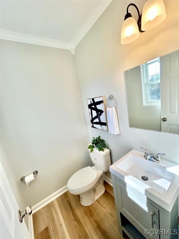 half bath with toilet, wood finished floors, vanity, baseboards, and ornamental molding