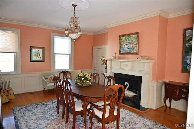 dining area with a wainscoted wall, a fireplace with raised hearth, wood finished floors, and crown molding