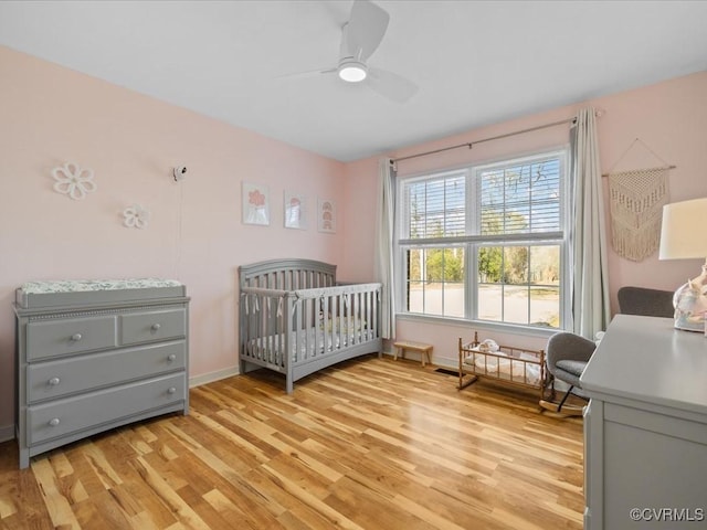 bedroom with a nursery area, light wood finished floors, a ceiling fan, and baseboards