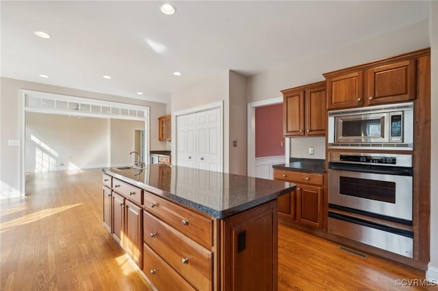 kitchen featuring light wood-type flooring, a kitchen island with sink, appliances with stainless steel finishes, and brown cabinetry