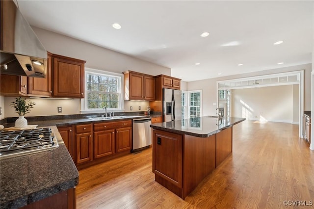 kitchen with light wood finished floors, wall chimney exhaust hood, appliances with stainless steel finishes, a center island, and a sink