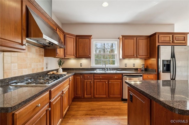 kitchen with stainless steel appliances, wood finished floors, a sink, wall chimney range hood, and brown cabinets