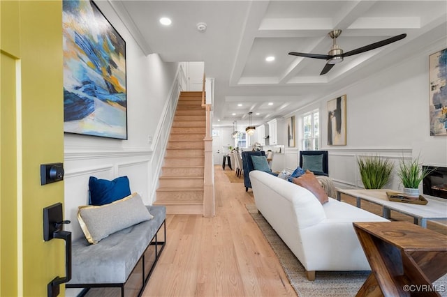 living area featuring beam ceiling, light wood finished floors, a decorative wall, coffered ceiling, and stairs