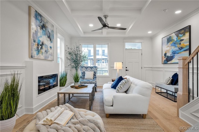 living area with crown molding, a decorative wall, a glass covered fireplace, light wood-type flooring, and coffered ceiling