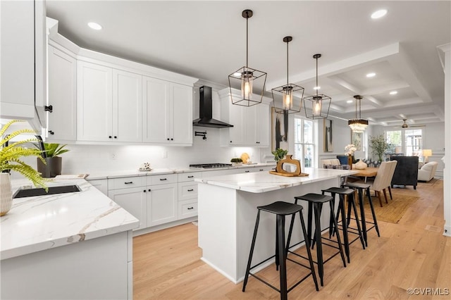 kitchen with light wood finished floors, coffered ceiling, wall chimney exhaust hood, a center island, and gas stovetop