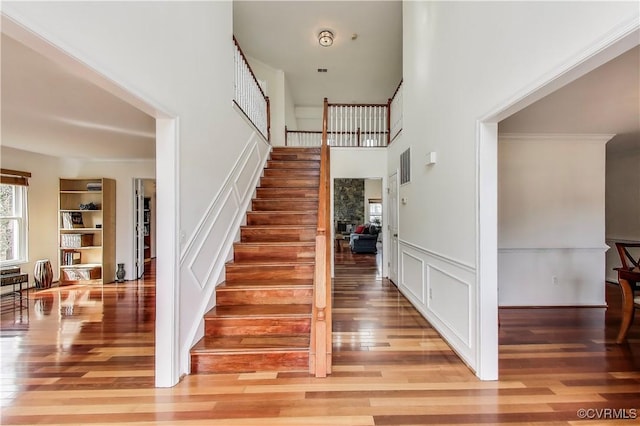 staircase featuring visible vents, wainscoting, wood finished floors, a high ceiling, and a decorative wall