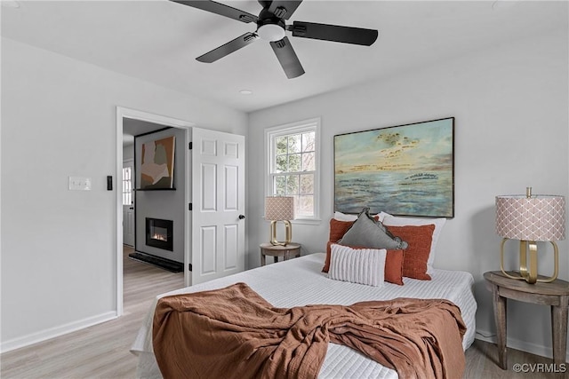 bedroom with a ceiling fan, a glass covered fireplace, baseboards, and light wood finished floors