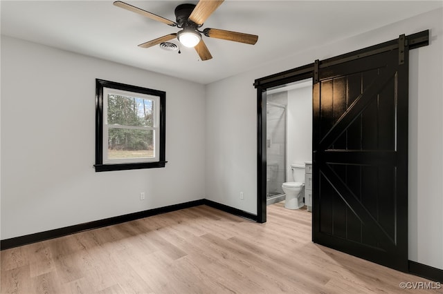 unfurnished bedroom with light wood finished floors, a barn door, baseboards, a ceiling fan, and connected bathroom