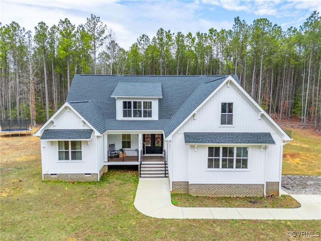 modern farmhouse style home with a front lawn, a trampoline, covered porch, a shingled roof, and crawl space