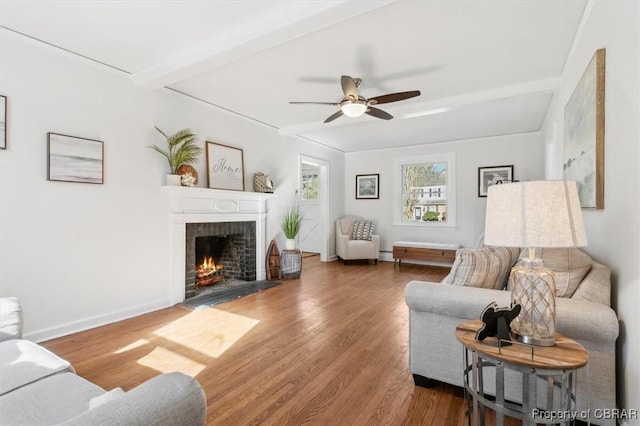 living room with ceiling fan, wood finished floors, baseboards, a brick fireplace, and beam ceiling