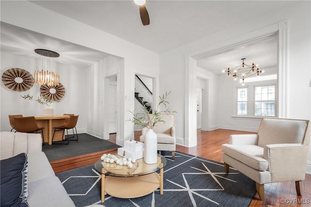 living area featuring ceiling fan with notable chandelier, stairway, wood finished floors, and baseboards