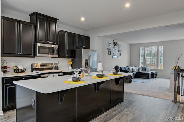 kitchen featuring open floor plan, light countertops, appliances with stainless steel finishes, dark cabinetry, and an island with sink