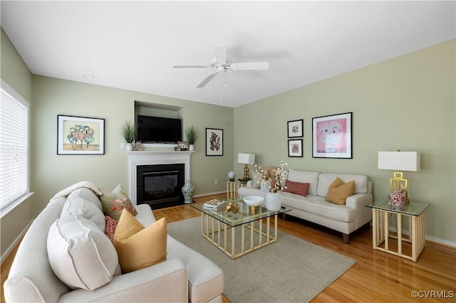 living room featuring a glass covered fireplace, ceiling fan, baseboards, and wood finished floors