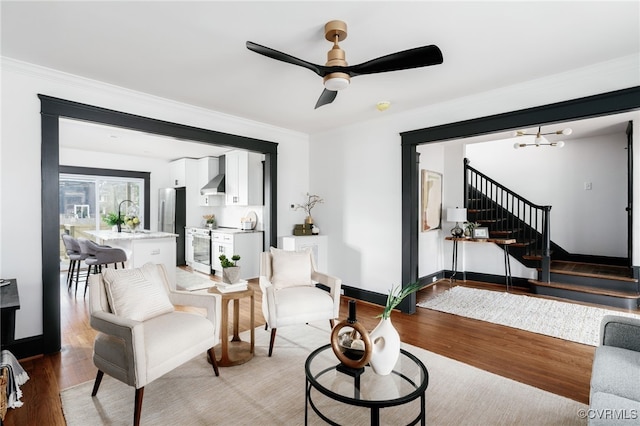 living room with baseboards, stairs, crown molding, light wood-style floors, and ceiling fan with notable chandelier